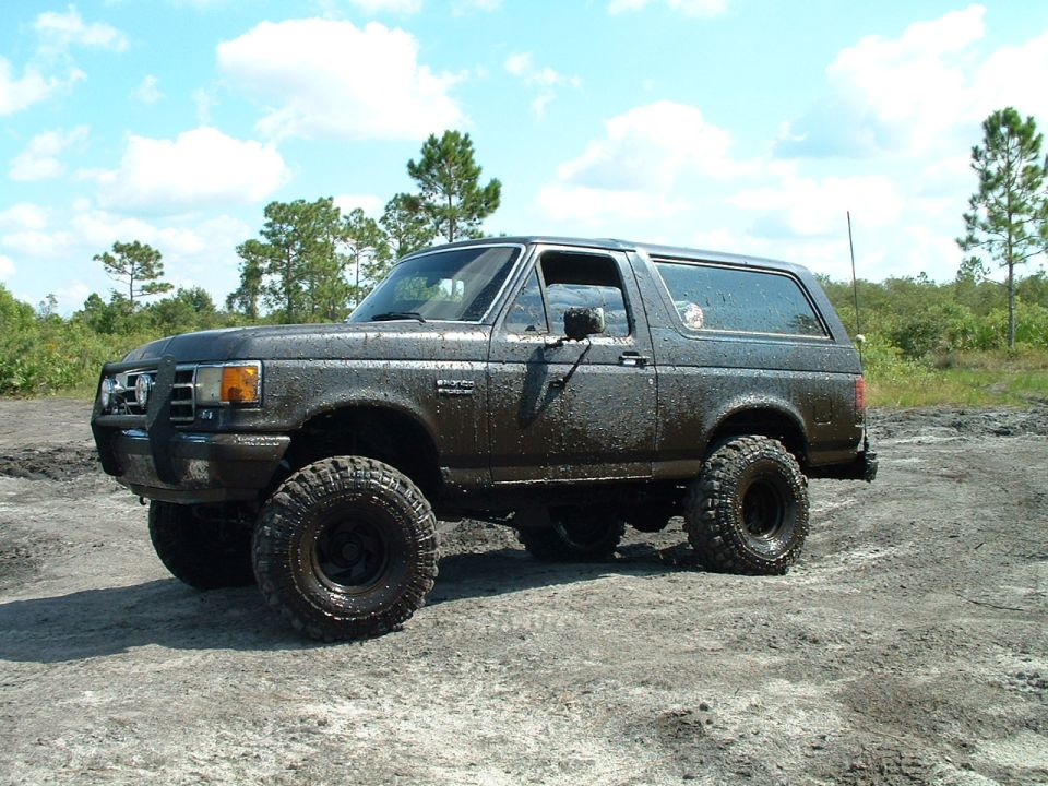 1995 Ford Bronco covered in mud, parked on a dirt track with trees in the background