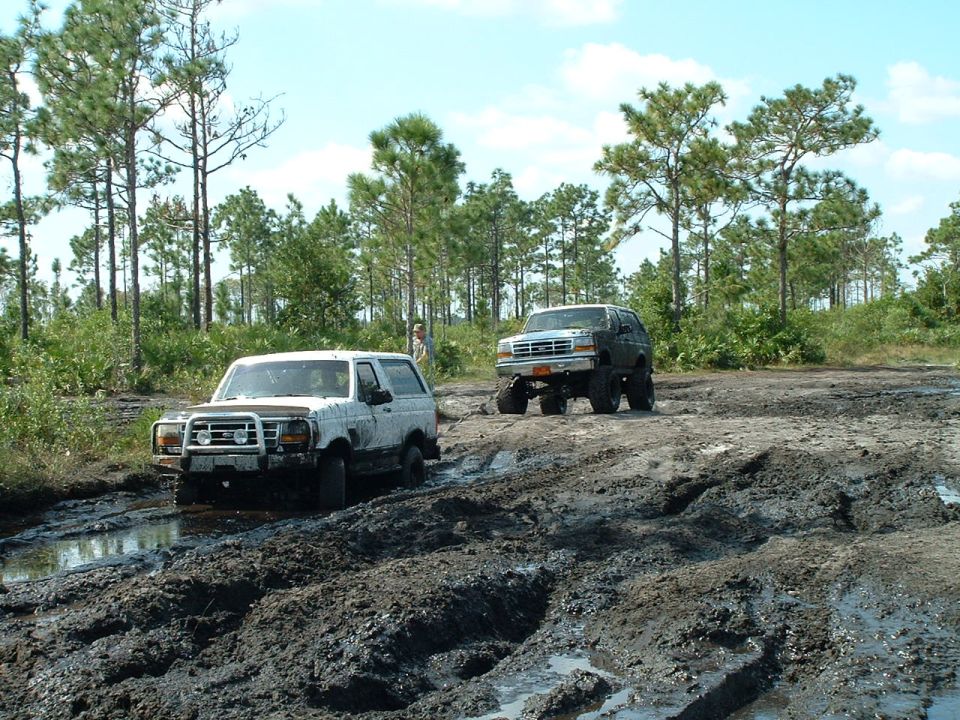 Two Ford Broncos driving through a muddy trail with trees in the background