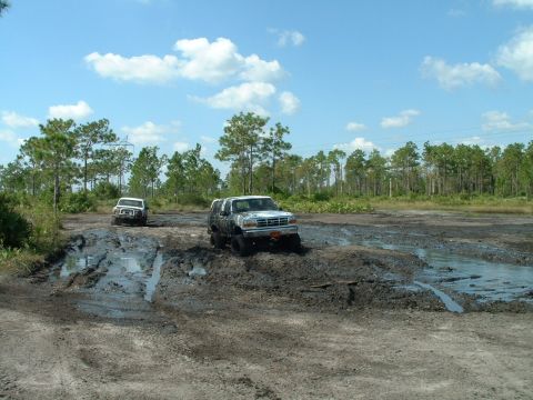 1995 Ford Bronco - Two SUVs navigate a muddy off-road course under a partly cloudy sky.