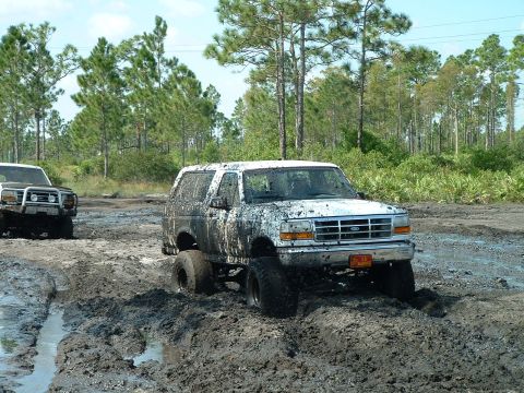 1995 Ford Bronco - Two SUVs navigate a muddy trail with trees in the background.