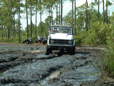 1995 Ford Bronco - A classic Ford Bronco navigates a muddy off-road course, with several ATVs and onlookers present.