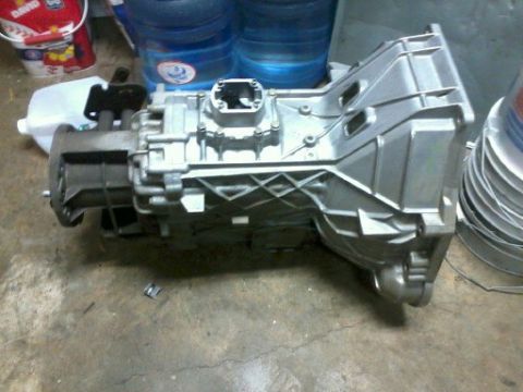 1985 Ford Bronco - A silver transmission part is shown on a concrete floor, with water jugs and a bucket in the background.