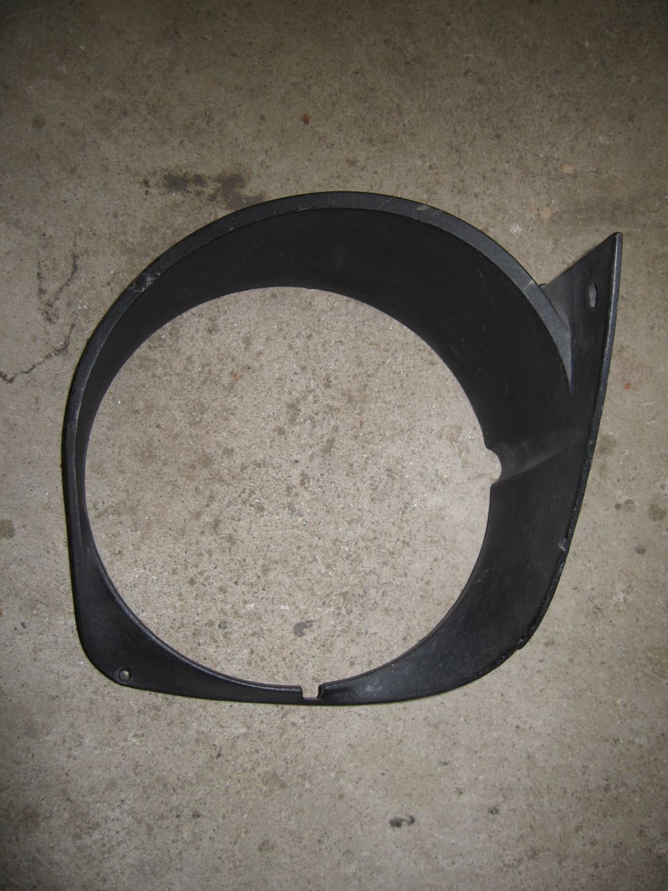 Black plastic headlight bezel with a triangular mounting tab, lying on a concrete surface