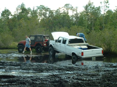 1995 Ford Bronco - A white Ford pickup truck, likely from the mid-1990s, is stuck in the mud with its hood open, while a red Jeep sits nearby.