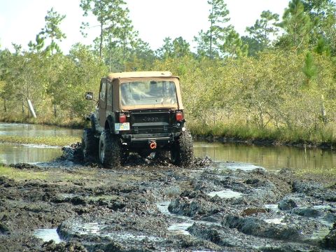 1995 Ford Bronco - A Jeep Wrangler navigates a muddy trail with trees and water in the background.