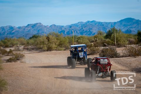 Suzuki Samurai - Two custom off-road vehicles traverse a dusty desert trail under a clear blue sky.