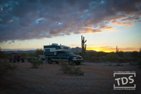 Suzuki Samurai - A truck camper is parked in a desert landscape with saguaro cacti under a dramatic sunset sky.