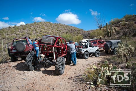 Suzuki Samurai - A group of modified off-road vehicles are gathered on a dusty trail in a desert environment.