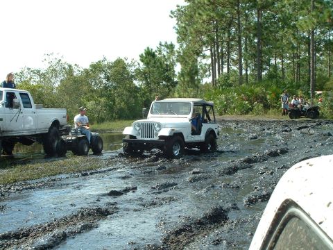 1995 Ford Bronco - Several off-road vehicles navigate a muddy track surrounded by trees and vegetation.