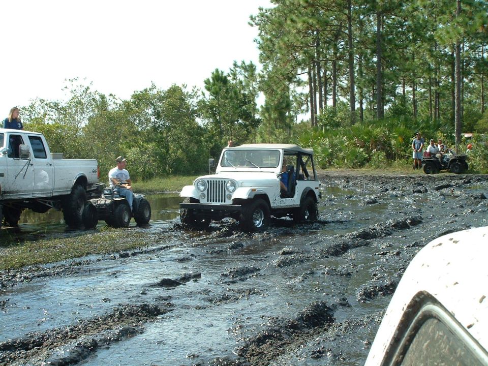 White Jeep Wrangler driving through mud, with a white pickup truck and an ATV in the background