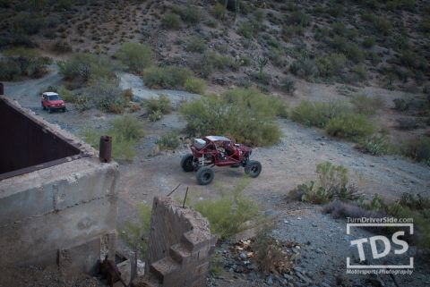 Suzuki Samurai - Two vehicles navigate a dusty desert trail near abandoned structures.