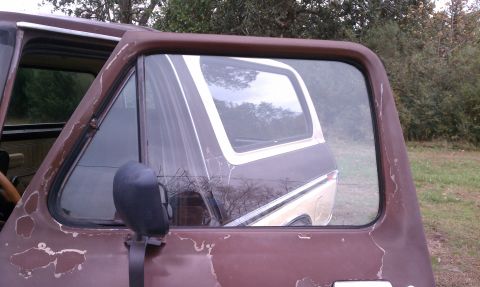 1979 Ford Bronco - Reflection in the window shows the rear of the vehicle. The paint is peeling on the door.