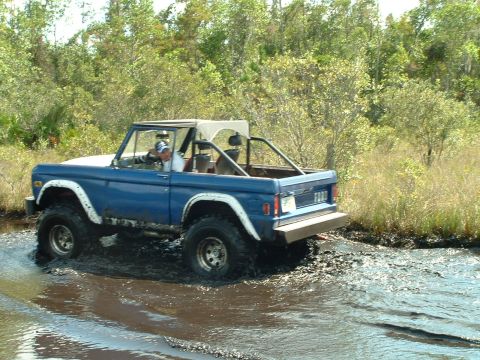 1995 Ford Bronco - A blue Ford Bronco navigates a muddy terrain with trees in the background.