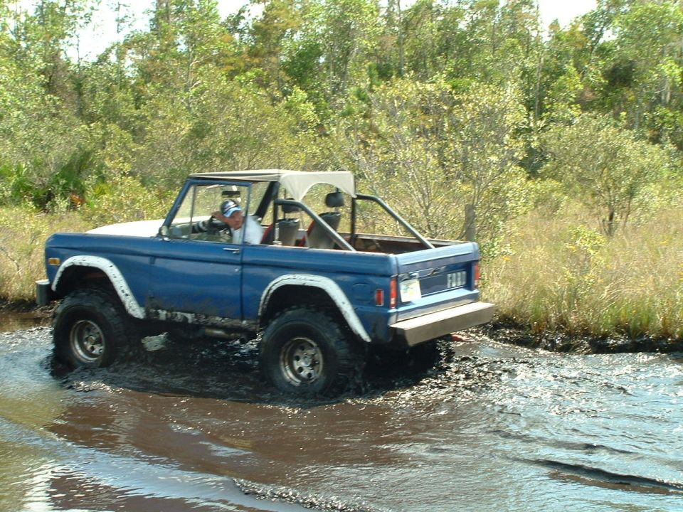 Blue Ford Bronco with white fender flares driving through mud, white soft top, roll cage