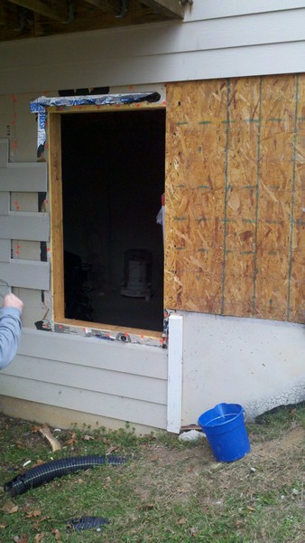 1995 Ford Bronco - Exterior wall under construction shows an open window frame and plywood sheeting, with a blue bucket and drainage pipe on the ground.