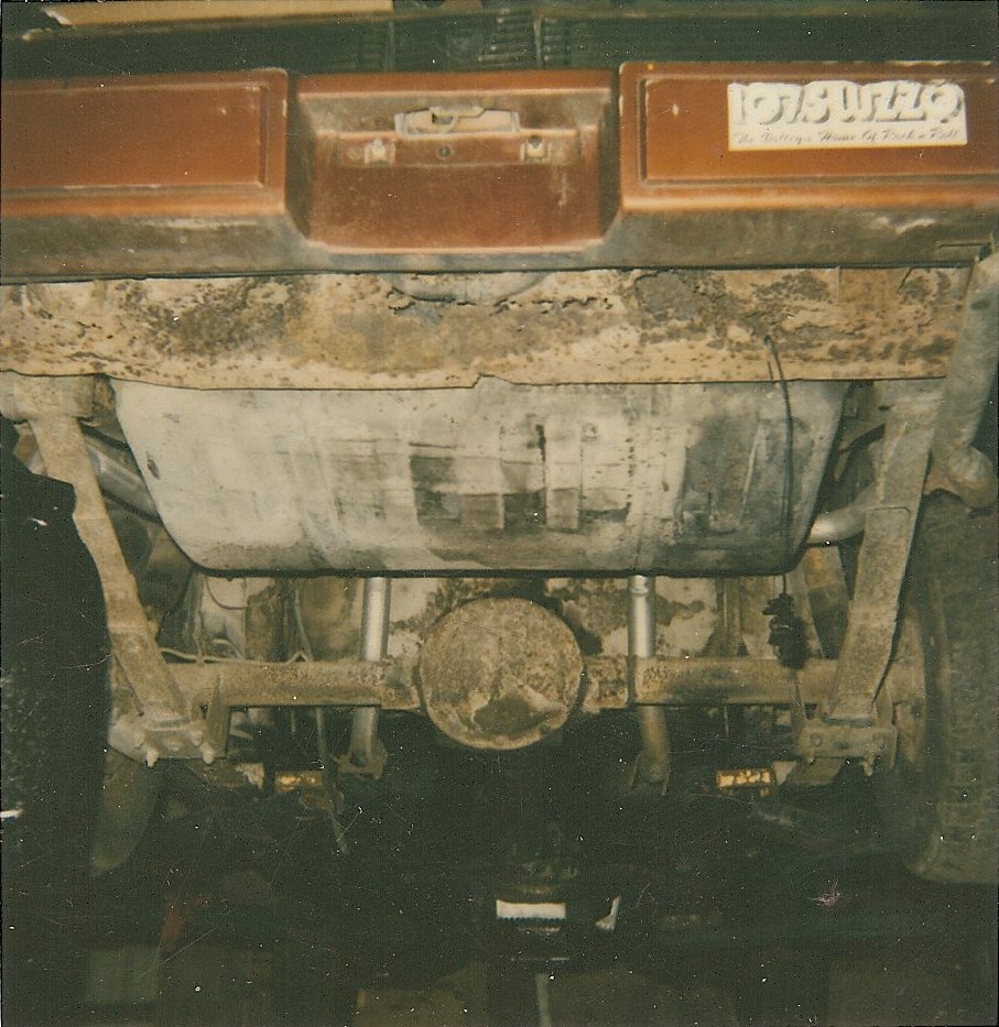 Underside view of a car's rear axle and fuel tank. A sticker on the bumper reads '107 BUZZO The Rolling Home Of Food & Fun'
