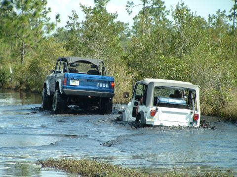 1995 Ford Bronco - Two off-road vehicles navigate through a water-filled terrain, with the Ford Bronco leading the way.