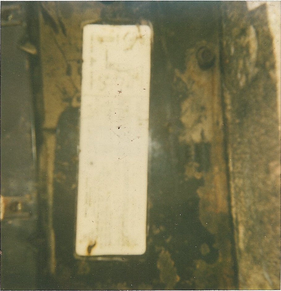 White rectangular plate with faint writing on a dark, dirty vehicle part
