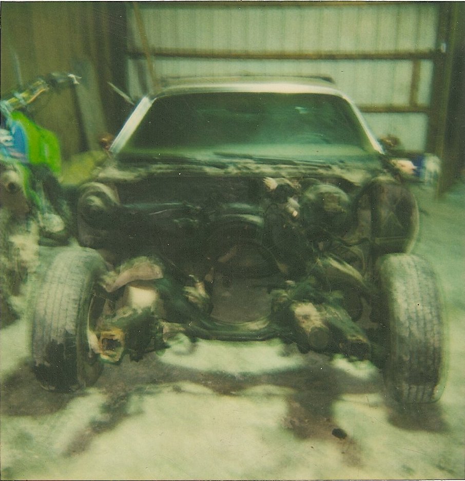 Front view of a disassembled car with engine exposed and tires on. A dirt bike is partially visible to the left