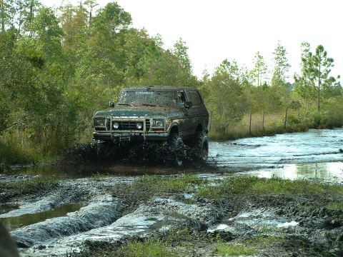 1995 Ford Bronco - A heavily muddied green Ford Bronco navigates a swampy terrain, kicking up water and mud.