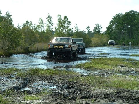 1995 Ford Bronco - Off-road vehicles navigate a challenging muddy water obstacle course under a bright sky.