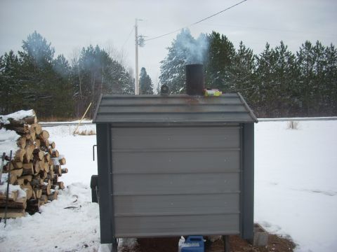 1963 Hummer H3 - A metal wood boiler is in operation, emitting smoke into the winter air, with a stack of firewood to its left.