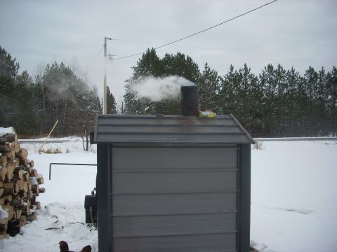 1963 Hummer H3 - A wood boiler emits smoke into the air on a cloudy, snowy day, with a stack of firewood nearby.