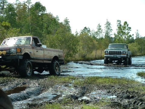 1995 Ford Bronco - Two pickup trucks navigate a challenging muddy course, with one truck in the foreground and another in the distance.