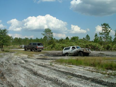 1995 Ford Bronco - Two SUVs navigate a muddy off-road course on a sunny day with scattered clouds.
