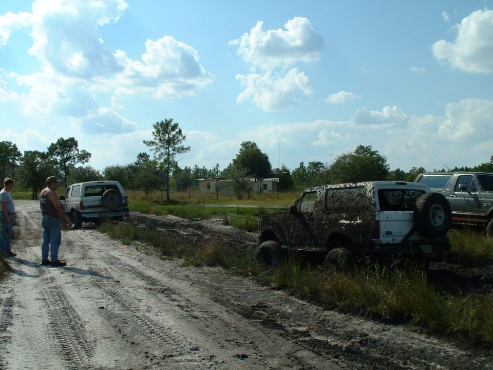 Mud-covered SUV stuck in mud, rear spare tire visible. Another white SUV and a pickup truck are nearby