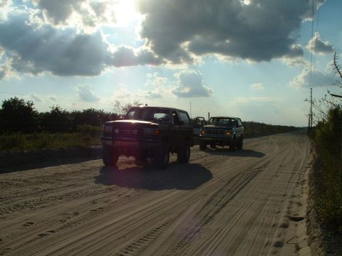 1995 Ford Bronco - Two SUVs travel down a dusty road, kicking up dust under a dramatic sky. The lead vehicle appears to be a Ford Bronco from the early 1990s.