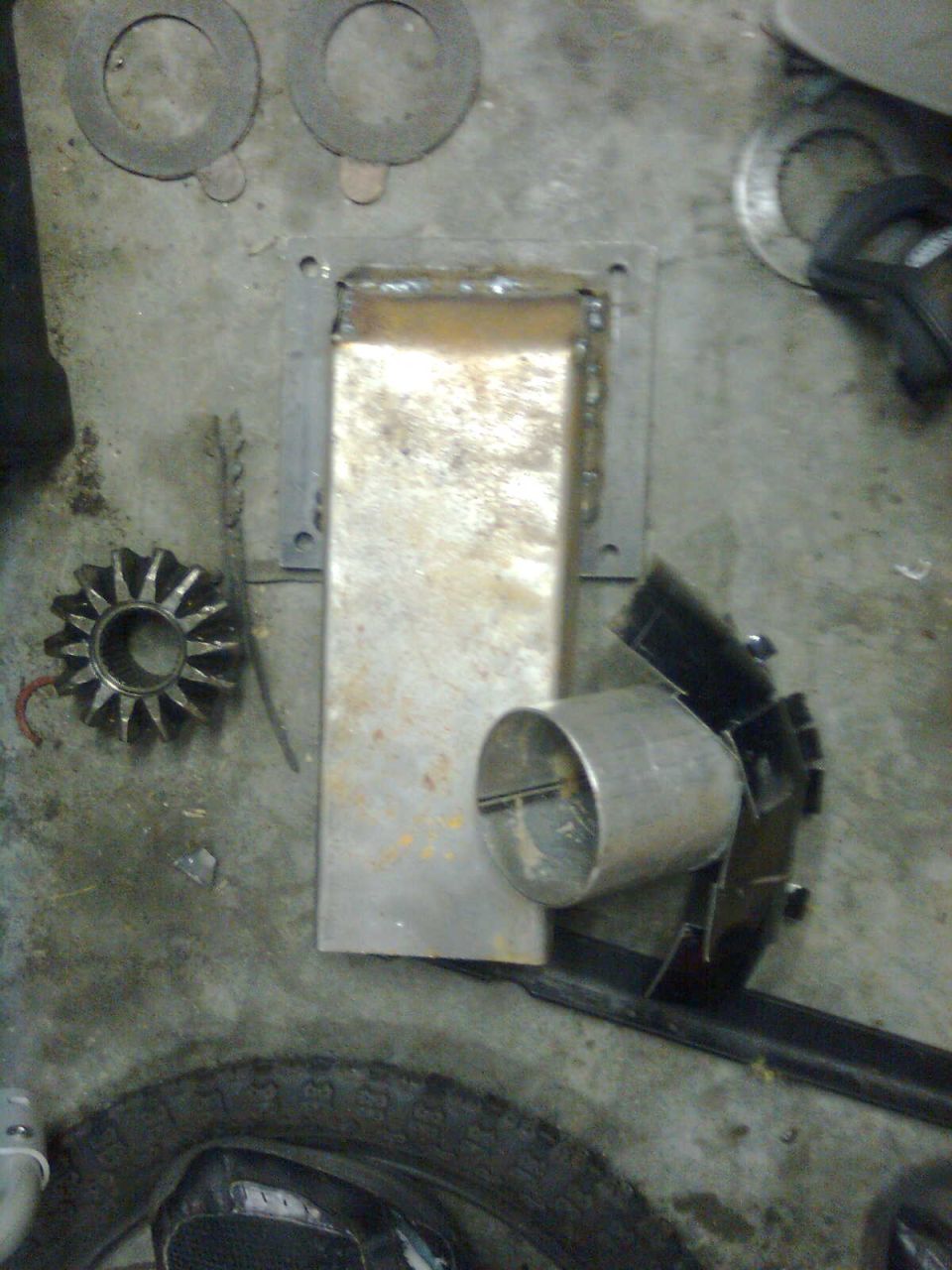 Metal plate with a welded rectangular opening, a cylindrical pipe, a gear-like part, and washers on a concrete floor next to a tire