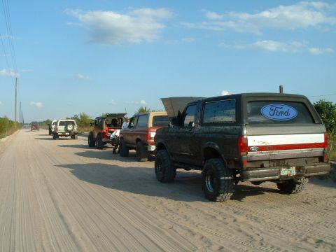 1995 Ford Bronco - Several Ford Broncos are lined up on a dirt road under a blue sky with scattered clouds.