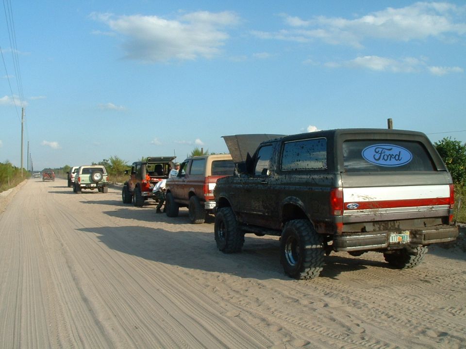 Line of lifted Ford Broncos on a dirt road, one with hood open. The closest Bronco has a Ford oval sticker on the rear window