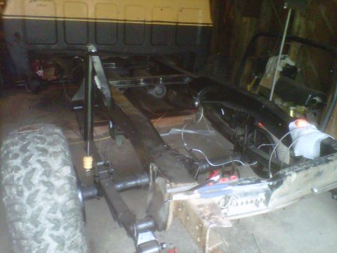 1978 Chevrolet C/K Pickup - The chassis of a truck is shown, with the frame, suspension, and a rear tire in focus. The cab is visible in the background.