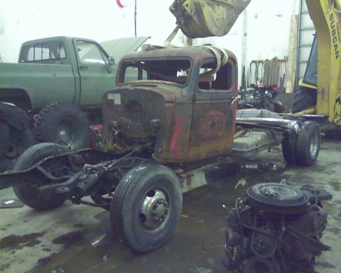 1978 Chevrolet C/K Pickup - A rusty truck chassis sits in a garage with a green pickup truck and a separate engine nearby.
