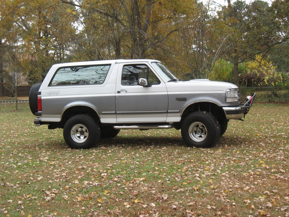 Two-tone white and silver Ford Bronco with a spare tire on the rear, parked on a grassy area with fallen leaves