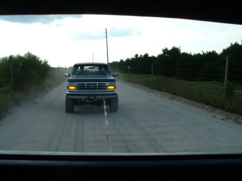 1995 Ford Bronco - A truck drives down a dusty road bordered by trees and a fence under a cloudy sky.