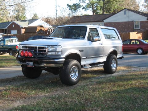 1994 Ford Bronco - A lifted, two-door Ford Bronco is parked on a grassy area next to a street with other vehicles visible.
