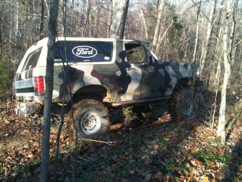 1995 Ford Bronco - A camouflaged Ford Bronco navigates a wooded terrain with its right side visible.