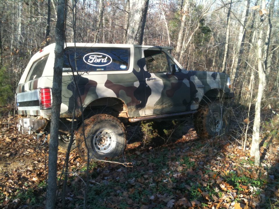 Camouflaged Ford Bronco with a white roof, seen from the right side in a wooded area
