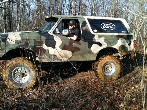 1995 Ford Bronco - Camouflaged 1995 Ford Bronco navigating a wooded trail with muddy tires.