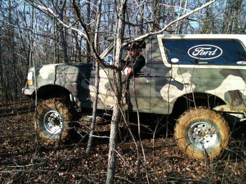 1995 Ford Bronco - A camouflage-painted Ford Bronco navigates through a wooded area with muddy tires.