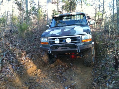 1995 Ford Bronco - This truck is navigating a muddy, wooded path, showing off its custom camouflage paint job and off-road bumper.