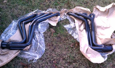 1979 Ford Bronco - Two sets of black exhaust headers are laid out on brown paper and plastic wrap on a grassy surface.