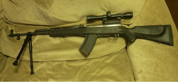 Rifle with scope and bipod resting on a tan couch
