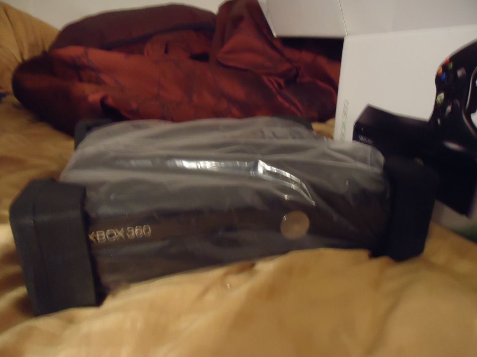 Xbox 360 console in plastic wrap, with a red blanket and a box in the background