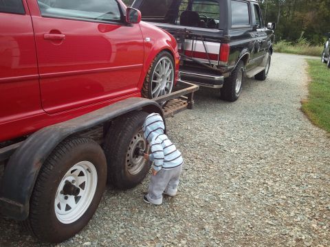 1989 Ford Bronco - A child inspects a trailer tire while a red car is loaded and a black Ford Bronco is parked behind.