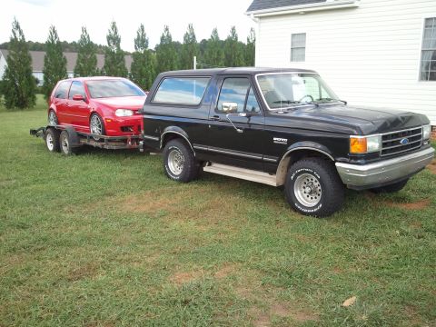 1989 Ford Bronco - A black Ford Bronco tows a red car on a trailer across a grassy yard.