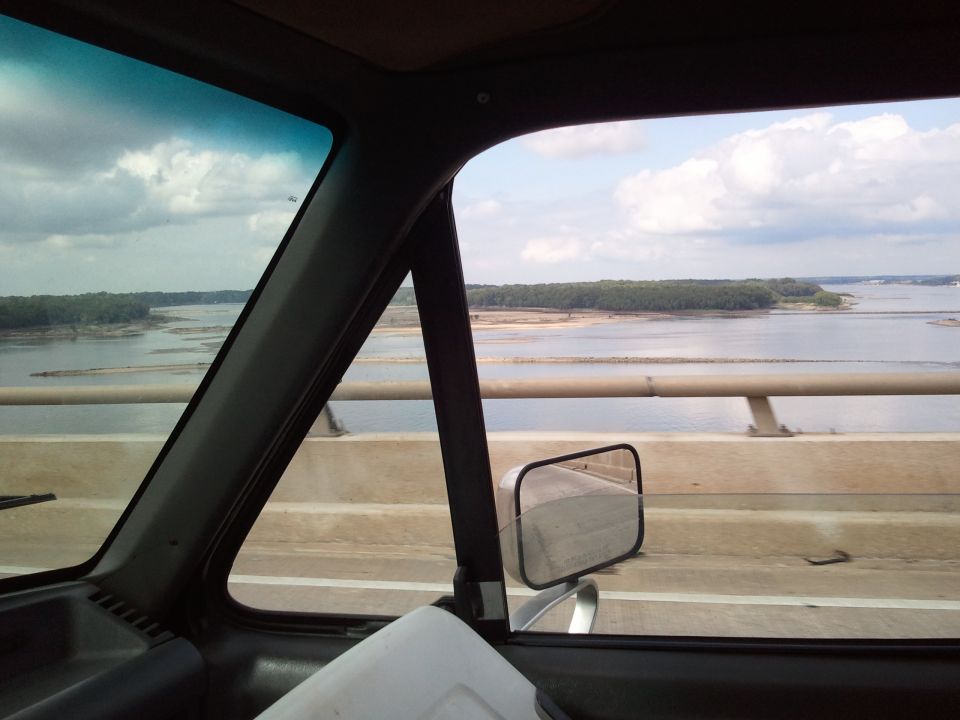 Interior view from a vehicle, looking out the passenger side window at a river and bridge. Watermark reads SUPERMOTORS